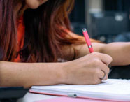 Aluna da Faculdade de Letras Inglês escrevendo em caderno com caneta rosa, em ambiente de estudo, focando em aprendizado e anotações.