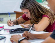 Alunos da Faculdade de Letras Inglês realizando atividade em sala de aula, escrevendo em cadernos, com foco em estudante com tatuagens no braço.