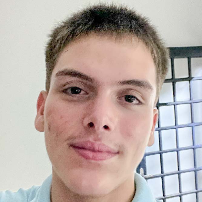 João Gabriel, aluno de Licenciatura em Educação Física, posando em um ambiente interno com uma grade ao fundo. Ele tem cabelos curtos e uma expressão neutra.