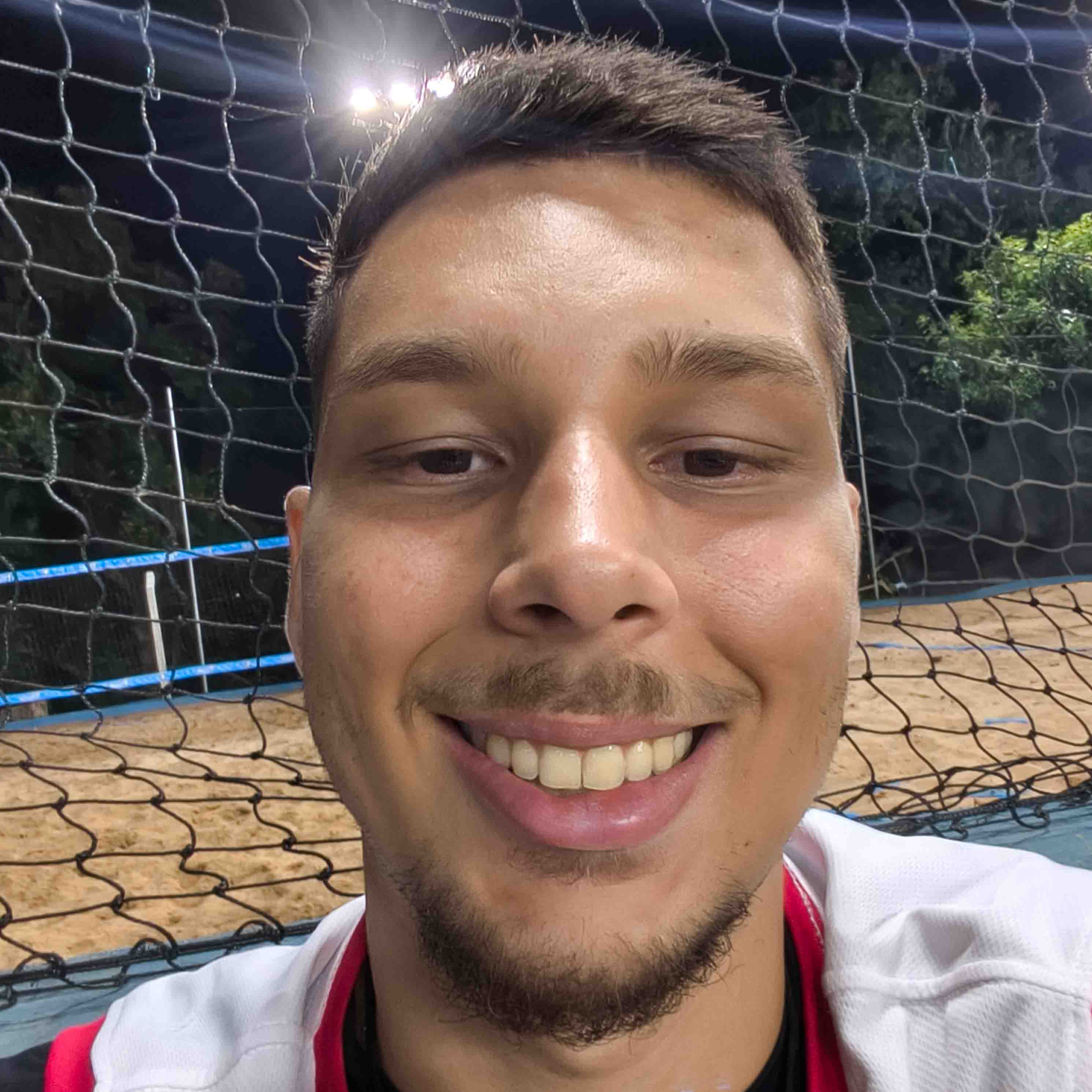 Lucas, egresso de Educação Física - Licenciatura, posando em um ambiente externo com uma rede de vôlei ao fundo. Ele tem barba rala e um sorriso largo.