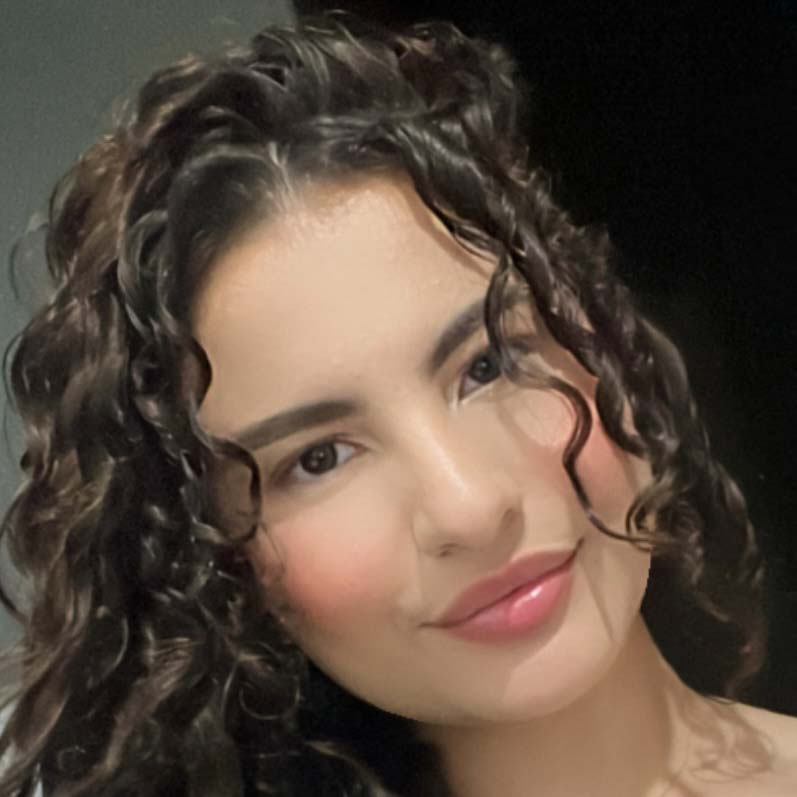 Maria Eugenia, aluna de Educação Física - Licenciatura, posando em um ambiente interno com um fundo escuro. Ela tem cabelos cacheados e um sorriso suave.