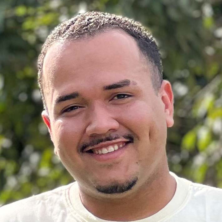 Victor, aluno de Educação Física - Licenciatura, posando em um ambiente externo com folhagens ao fundo. Ele tem barba rala e um sorriso largo.