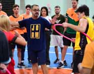 Turma do curso de Educação Física da Unoeste reunida na quadra, com destaque para professor de camiseta azul JOAM orientando estudantes. Aluno de colete amarelo segura bambolê rosa, enquanto outros observam. Estudantes vestem coletes laranja e uniformes esportivos, formando um círculo de atenção à explicação.