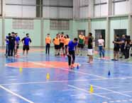 Estudantes do curso de Educação Física da Unoeste realizam gincana na quadra coberta. Participantes com coletes amarelos, azuis e laranja aguardam em fila, enquanto outros conduzem bambolês rosas entre cones coloridos. Professores acompanham e orientam a atividade, promovendo integração e prática pedagógica.