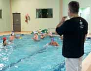 Aula prática de natação do curso de Educação Física da Unoeste, campus Presidente Prudente. Estudantes dentro da piscina usam toucas coloridas e praticam exercícios aquáticos, supervisionados por professor à beira da piscina com apito. Equipamentos de natação e boias estão ao fundo, em ambiente fechado.