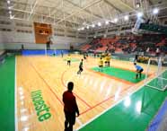 Ginásio poliesportivo da Unoeste, campus Presidente Prudente, com quadra de piso claro, demarcações verdes e vermelhas, e arquibancadas laranja. Partida de futsal em andamento, jogadores de uniforme verde e amarelo, torcida acompanhando nas arquibancadas. Estrutura moderna, iluminação forte e redes de proteção.