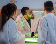 Professor da Faculdade de Celulose e Papel explicando conceitos de química para alunos em laboratório, com equipamentos de pesquisa.