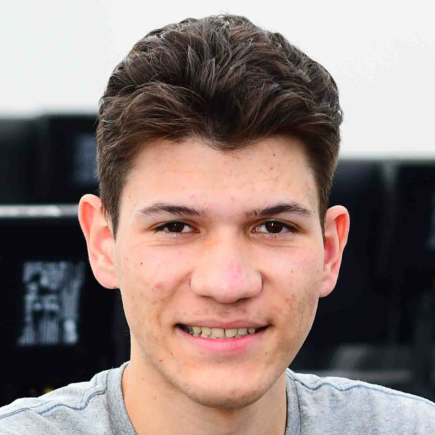 Ademir, aluno do curso de sistemas de informação, trajando uma camiseta cinza. Ele está em um ambiente interno com fundo composto por computadores e equipamentos tecnológicos, sugerindo um laboratório ou sala de informática. A iluminação clara destaca os detalhes da vestimenta e reforça o contexto acadêmico e tecnológico.