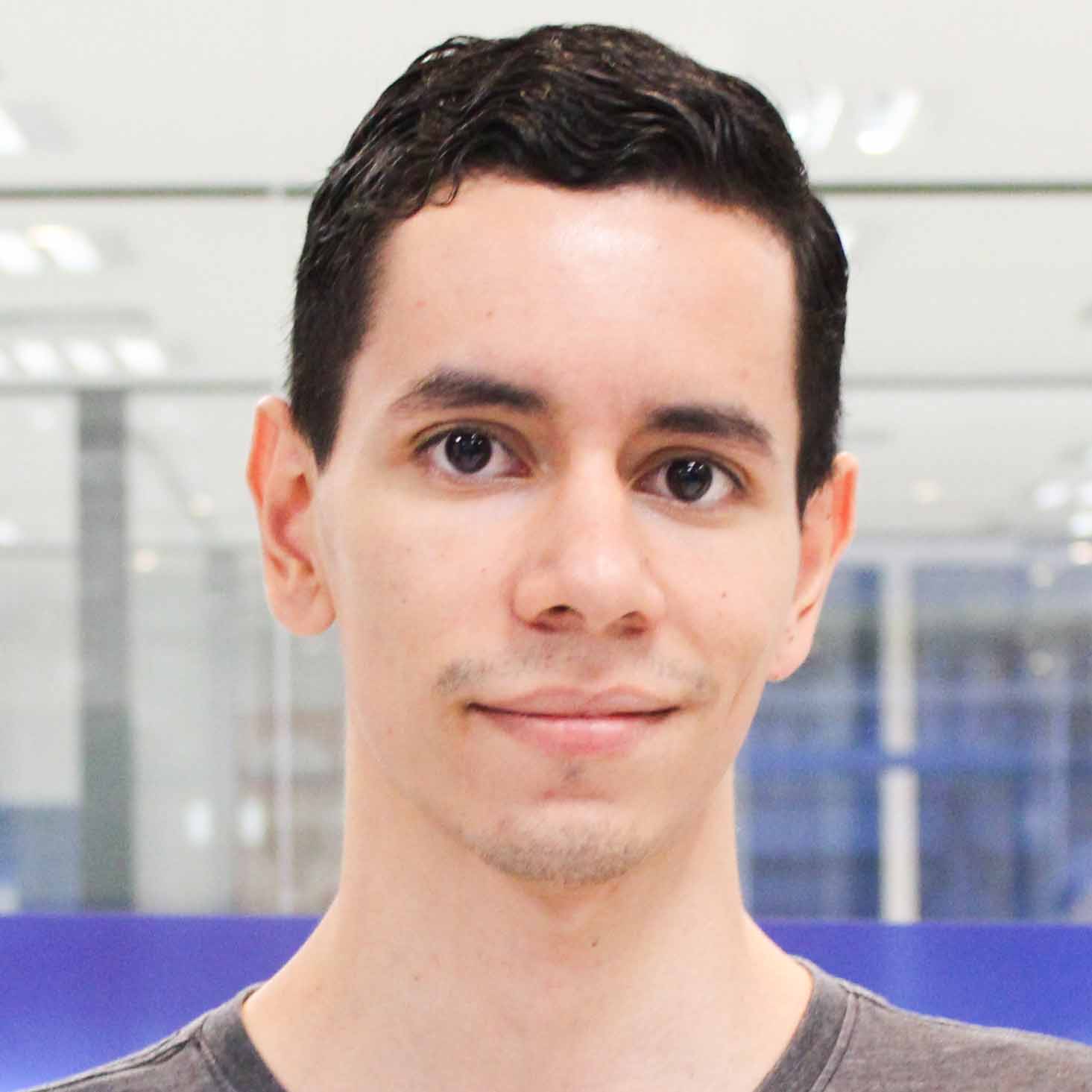 Bruno, egresso do curso de sistemas de informação, trajando uma camiseta cinza. Ele está em um ambiente interno com fundo moderno composto por paredes de vidro e tons azulados, indicando um espaço corporativo ou institucional. A iluminação uniforme valoriza a composição profissional e tecnológica da imagem.