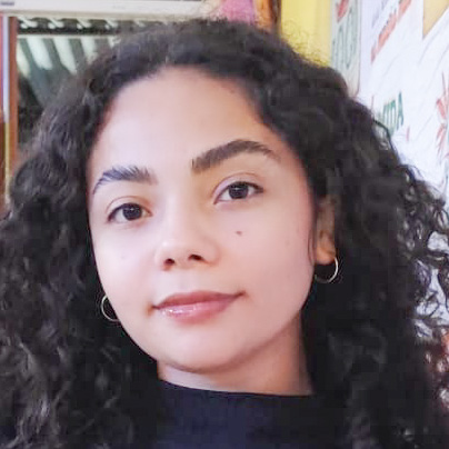 Mary, aluna do curso de sistemas de informação, trajando uma blusa preta. Ela está em um ambiente interno com fundo decorado por cartazes e elementos coloridos, sugerindo um espaço descontraído ou cultural. A iluminação natural destaca os cabelos cacheados e cria uma atmosfera acolhedora e dinâmica.