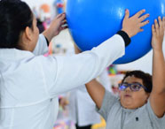 Aluna do curso de Terapia Ocupacional auxiliando paciente infantil em exercício de reabilitação com bola azul, demonstrando prática clínica e cuidado.