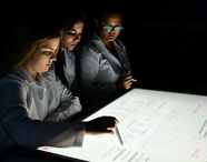 Professor e alunos da Faculdade de Terapia Ocupacional em Guarujá em aula na Sala Betha, utilizando mesas interativas para aprendizado tecnológico e colaborativo.