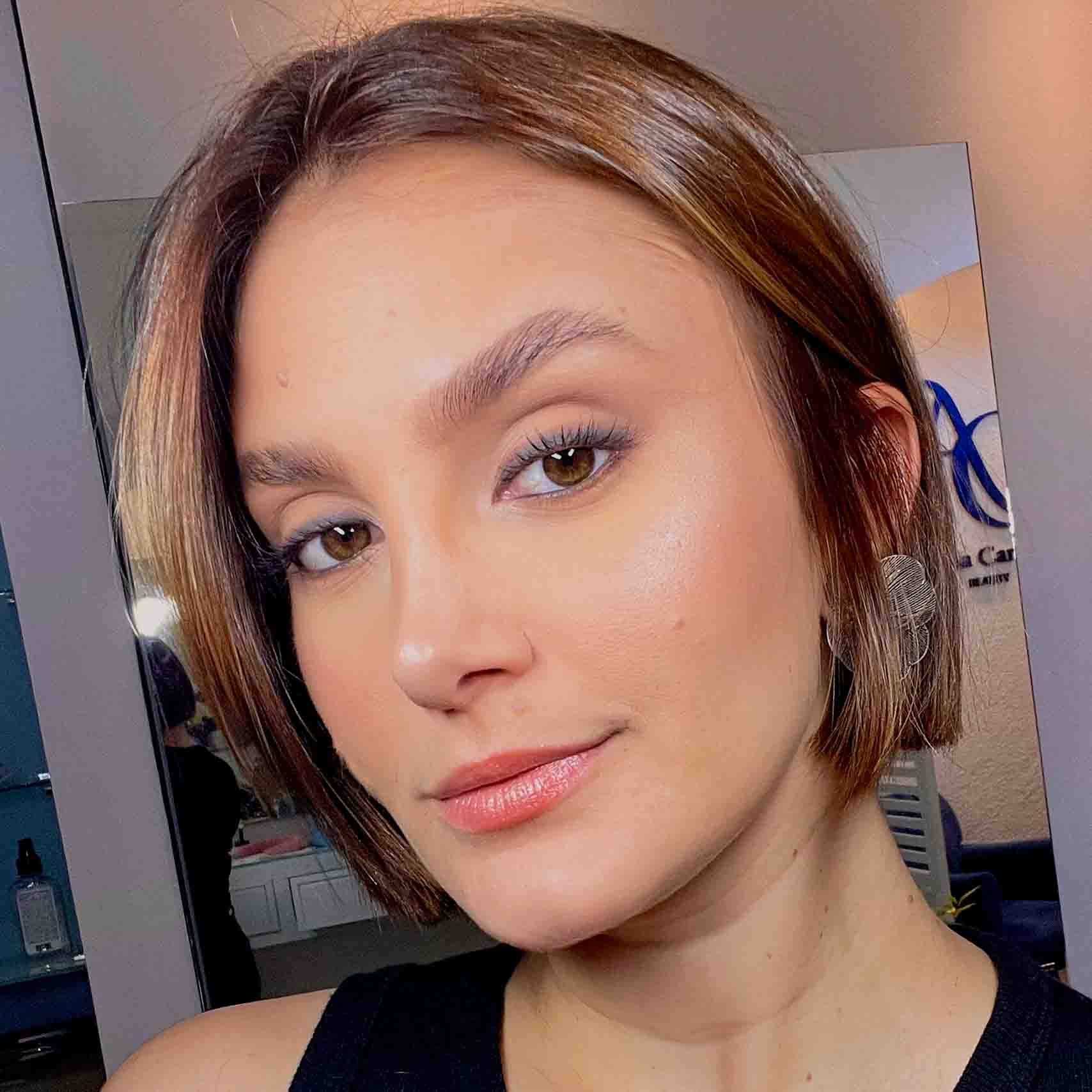 Ângela, aluna do curso de estética e cosmética, em um ambiente interno com espelhos e iluminação suave ao fundo. Ela veste uma blusa preta e acessórios discretos.