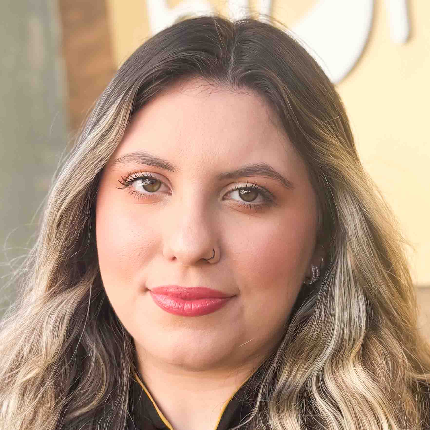 Gabriela, aluna do curso de estética e cosmética, em um ambiente externo com tons quentes ao fundo. Seu cabelo ondulado com mechas claras compõe a cena com elegância.