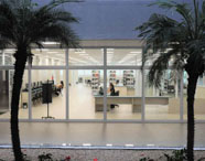 Vista interna da biblioteca da Faculdade de Estética e Cosmética em Guarujá, com grandes janelas, mesas de estudo e prateleiras de livros.
