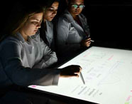 Alunas da Faculdade de Estética e Cosmética em Guarujá em aula na Sala Betha, utilizando mesas interativas para estudos avançados em ambiente tecnológico.