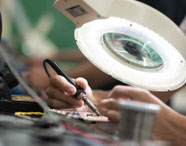 Aluno da Faculdade de Energias Renováveis em aula prática, realizando soldagem em placa eletrônica sob lupa iluminada, focando em eletrônica e sistemas.