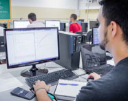 Aluno da Faculdade de Energias Renováveis em sala de aula, utilizando computador para estudos e projetos, com professor ao fundo.