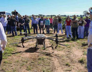 Drone agrícola realizando pulverização em lavoura, observado por estudantes e profissionais da Faculdade de Agroecologia em Presidente Prudente, destacando tecnologia no campo.