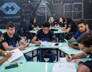 Faculdade de administração em Presidente Prudente, atividade em grupo sala de metodologias: estudantes sentados em mesas formam grupos de estudo, com livros, cadernos e celulares, enquanto conversam e resolvem atividades. O ambiente descontraído e colaborativo, com quadro-negro repleto de conteúdos, incentiva o protagonismo e a aprendizagem ativa em administração.