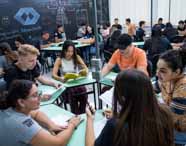 Faculdade de administração em Presidente Prudente, aula sala de metodologias ativas: estudantes divididos em grupos ocupam mesas verdes, interagindo e trocando ideias durante a aula. O quadro-negro ao fundo está repleto de anotações. O ambiente moderno e dinâmico valoriza o protagonismo discente, a construção coletiva do conhecimento e o desenvolvimento de habilidades essenciais para o futuro administrador.