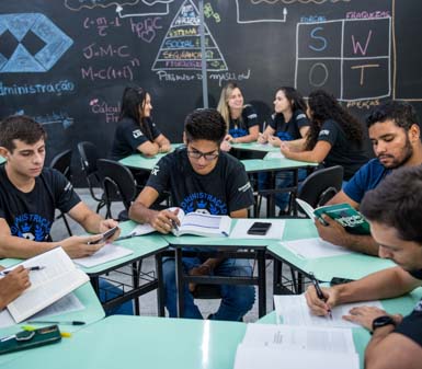 Estudantes do curso de Administração no campus Presidente Prudente, usando camisetas da graduação, estão reunidos em grupos ao redor de mesas, realizando atividades de estudo e pesquisa. Ao fundo, quadro negro com anotações de conceitos como SWOT, gráficos e fórmulas, reforçando o clima dinâmico, colaborativo e acadêmico da sala de aula, voltado ao desenvolvimento profissional.