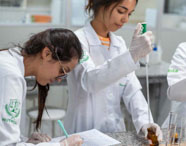 Alunas da Faculdade de Alimentos realizando análise química em laboratório, manuseando pipetas e reagentes para estudos de composição alimentar.