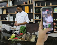 Chef da Faculdade de Alimentos realizando demonstração culinária em laboratório, sendo filmado por aluno, combinando aula prática e produção de conteúdo.