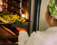 Aluna da Faculdade de Alimentos colocando bandeja de alimentos no forno em laboratório de gastronomia, demonstrando técnicas de preparo e cocção.