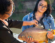 Aluna da Faculdade de Aquicultura segurando um peixe pacu, enquanto professora aponta para detalhes, em aula prática de piscicultura.