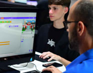 Aluno da Faculdade de Sistemas de Informação aprendendo programação em computador, com instrutor auxiliando na tela de código de blocos.