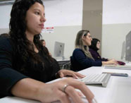 Aluna da Faculdade de Mídias Sociais Digitais concentrada em atividade no computador, utilizando mouse e teclado em laboratório de informática.