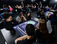 Alunos da Faculdade de Mídias Sociais Digitais em aula na Sala Betha, utilizando mesas interativas para aprendizado colaborativo e tecnológico.