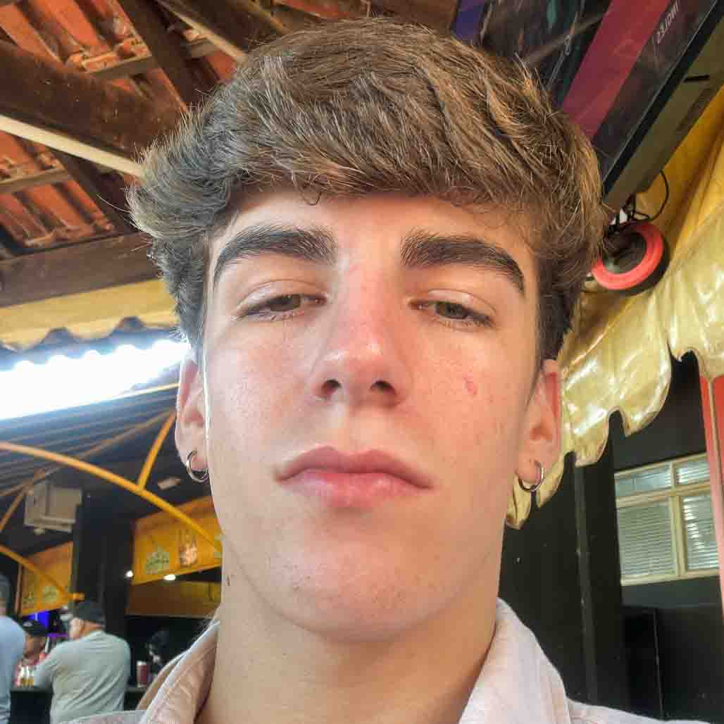 Cauê, aluno do curso de ciências biológicas (bacharelado), em um ambiente externo com estrutura de madeira e detalhes de toldos amarelos ao fundo. A iluminação natural destaca o cenário descontraído.