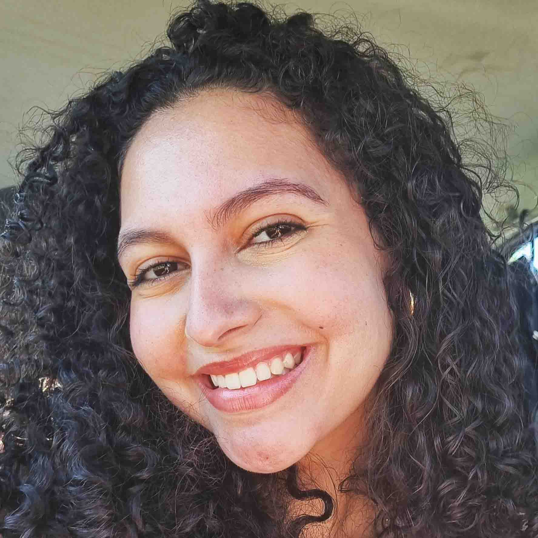 Débora, aluna do curso de ciências biológicas (bacharelado), em um ambiente interno com fundo neutro. Seu cabelo cacheado escuro compõe a cena com textura marcante.