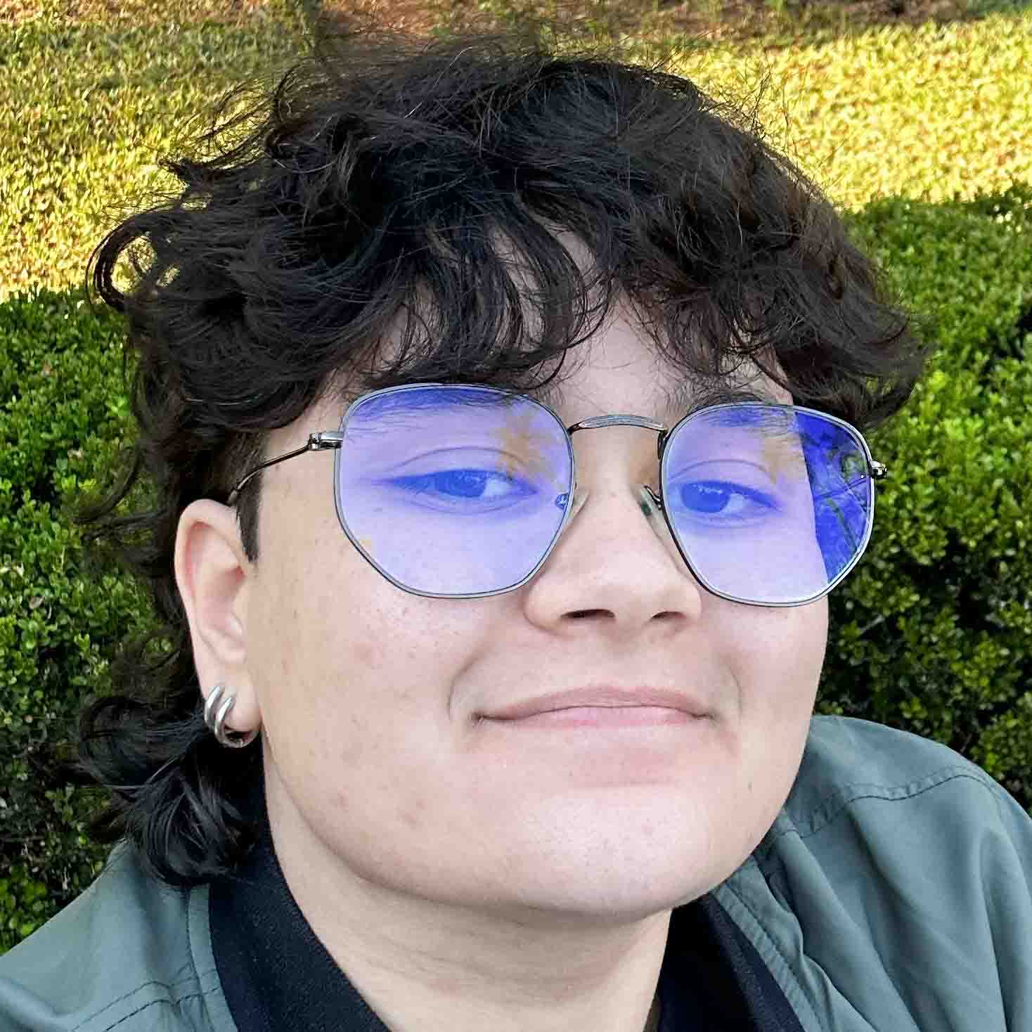 Oliver, aluno do curso de ciências biológicas (bacharelado), em um ambiente externo com arbustos verdes ao fundo. Ele usa óculos com lentes coloridas e veste uma jaqueta cinza, compondo um visual casual.