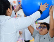 Aluna do curso de Terapia Ocupacional em Jaú auxiliando paciente infantil em exercício de reabilitação com bola azul, demonstrando prática clínica.