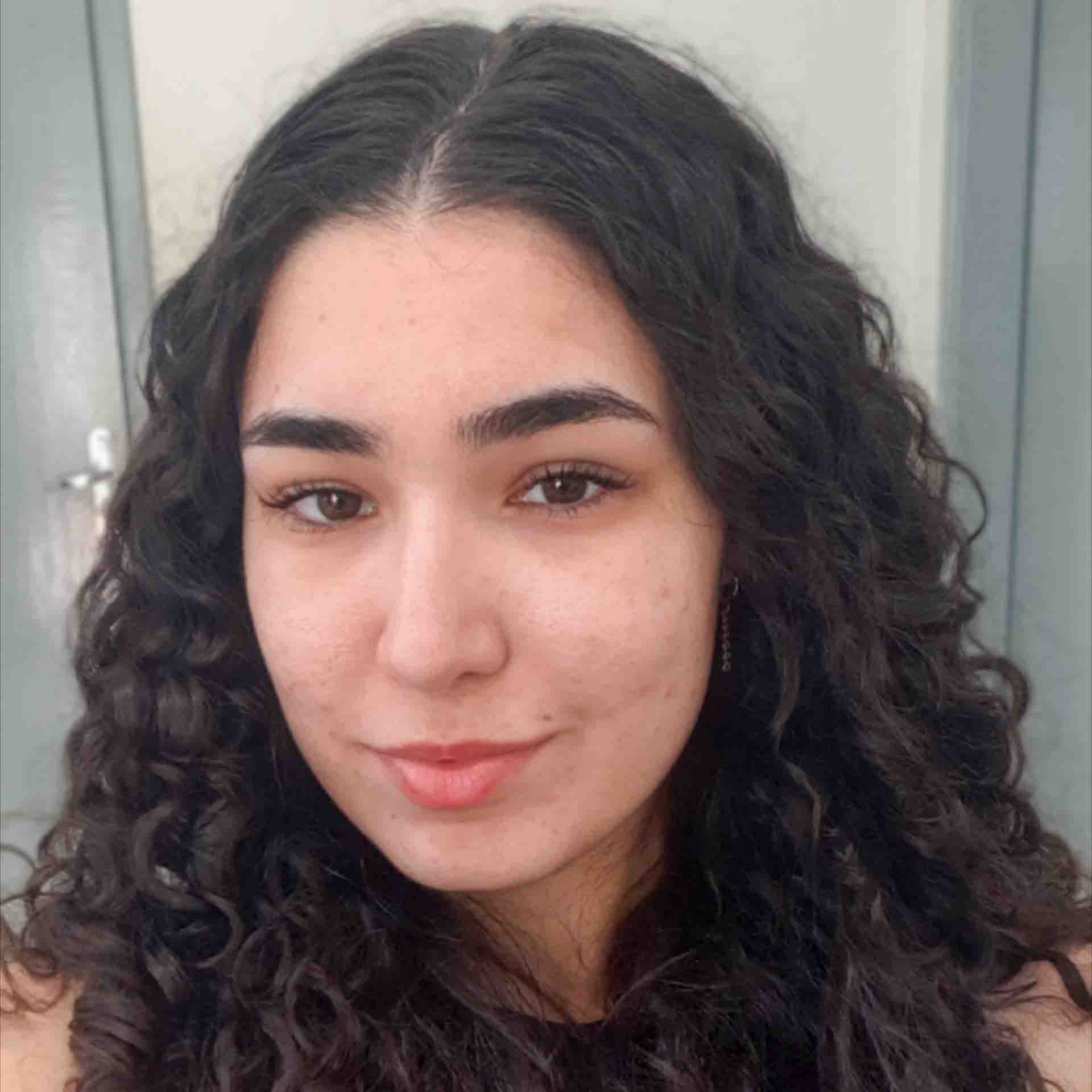 Giovanna, aluna do curso de ciências contábeis, posando em um ambiente interno com portas cinzas ao fundo. Seu cabelo cacheado escuro adiciona textura à composição.
