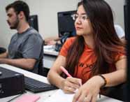 Estudantes do curso de Ciências Contábeis da Unoeste em Presidente Prudente participam de aula em laboratório de informática. Uma aluna de cabelos longos e blusa laranja faz anotações, enquanto outros alunos trabalham nos computadores ao redor, em ambiente colaborativo.
