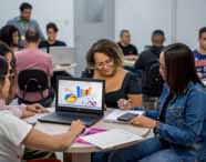 Grupo de estudantes do curso de Ciências Contábeis da Unoeste em Presidente Prudente reúne-se em torno de mesa redonda. No centro, notebook exibe gráficos coloridos. Alunas usam roupas casuais, uma delas com jaqueta jeans, todas envolvidas em discussão sobre análise de dados.