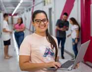 Estudante do curso de Ciências Contábeis da Unoeste segura notebook aberto no corredor da escola de negócios. Ela veste camiseta clara com estampa de borboleta. Ao fundo, grupo de colegas conversa próximo às paredes brancas e detalhes em rosa, transmitindo clima universitário.