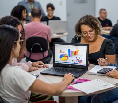 Estudantes do curso de Ciências Contábeis em Presidente Prudente estão reunidos em uma sala de aula, sentados ao redor de uma mesa redonda. Sobre a mesa há um notebook exibindo gráficos coloridos de barras e pizza, além de calculadora, papéis e canetas, indicando análise de dados financeiros e trabalho em grupo. Ao fundo, outros alunos utilizam notebooks em ambiente colaborativo.