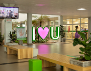 Saguão de entrada da Unoeste Jaú com bancos de madeira, plantas decorativas e um letreiro luminoso com a mensagem 'I ♡ U'.