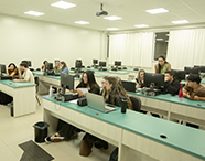 Alunos e professora do curso de Direito interagindo e realizando atividades em um laboratório de informática equipado com computadores desktop e notebooks.
