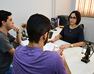Professora de Direito orientando dois alunos em sua mesa de atendimento com documentos e a estátua da Justiça.