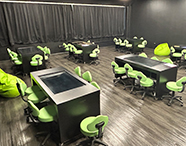 Vista ampla da Sala Betha Unoeste, um laboratório de aprendizagem com paredes escuras, mesas com monitores e cadeiras verdes ergonômicas.