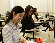 Estudantes universitárias da Faculdade de Direito sentadas em carteiras de sala de aula, sorridentes e concentradas, utilizando tablets e cadernos para anotações.