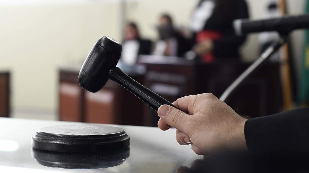 Mão segurando um martelo de juiz de madeira preta apoiado sobre o cepo, com o ambiente do tribunal desfocado ao fundo.