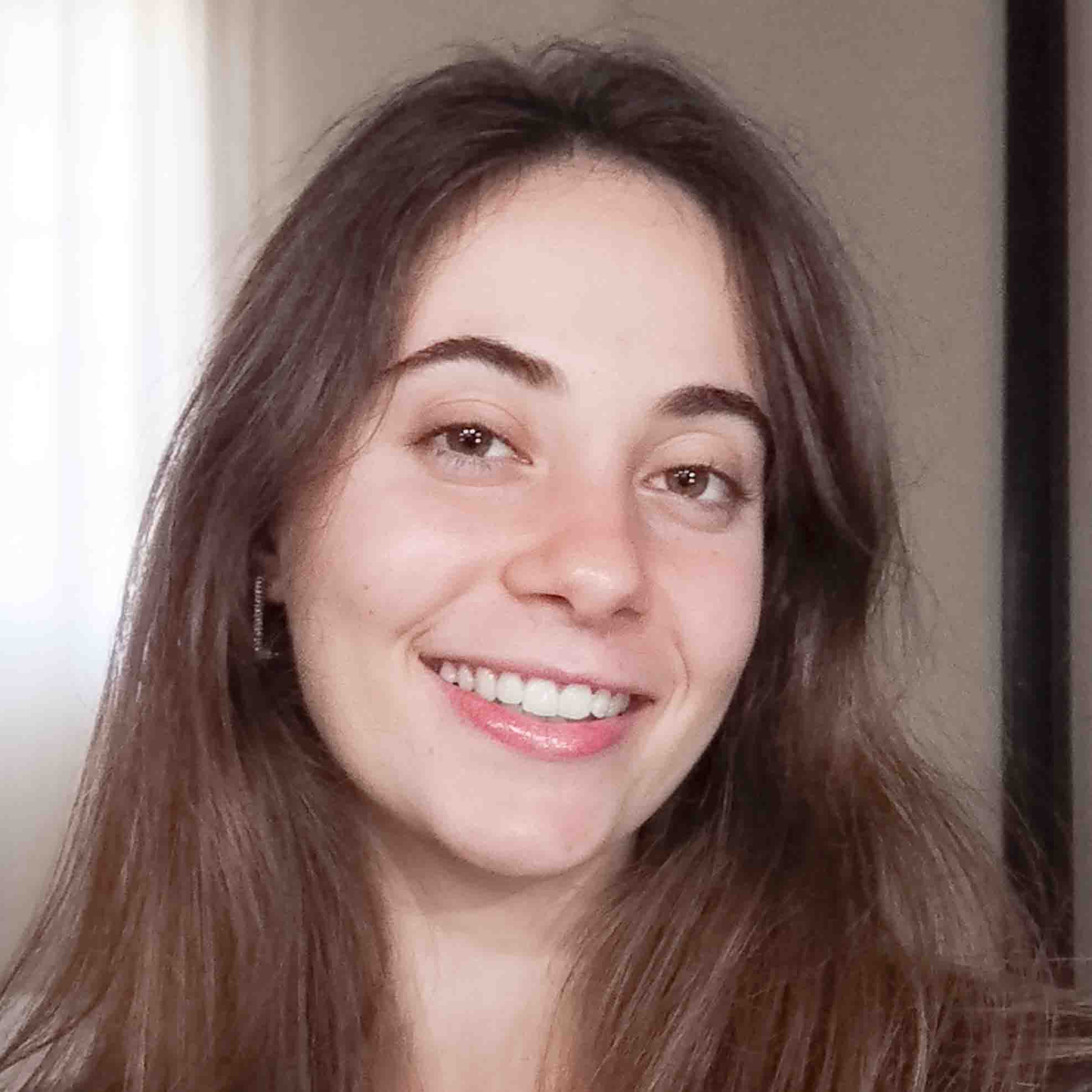 Juliana, aluna de Nutrição, posando em um ambiente interno com uma janela ao fundo. Ela tem cabelos longos e lisos, e usa brincos pequenos, com um sorriso largo.