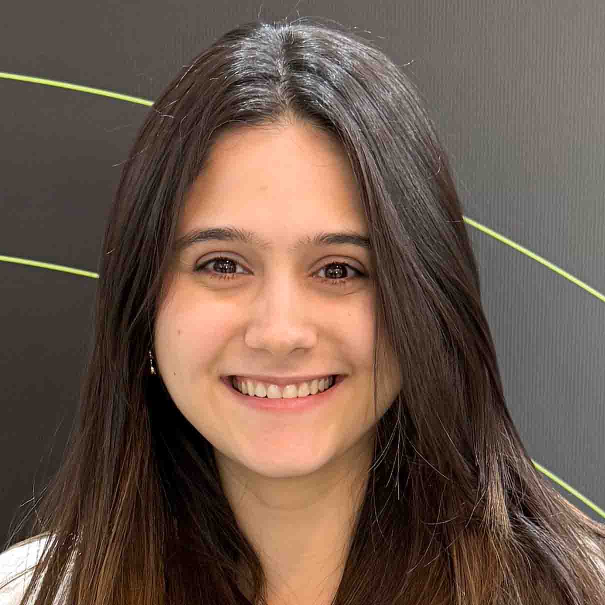Mariana, aluna de Nutrição, posando em um ambiente interno com um fundo escuro e linhas amarelas. Ela tem cabelos longos e lisos, e usa brincos pequenos, com um sorriso largo.